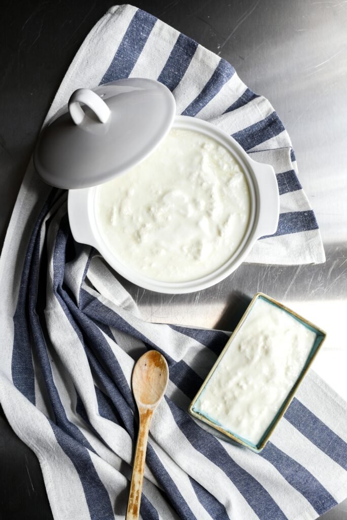 Top view of homemade yogurt in a ceramic pot, styled with a wooden spoon and striped cloth.