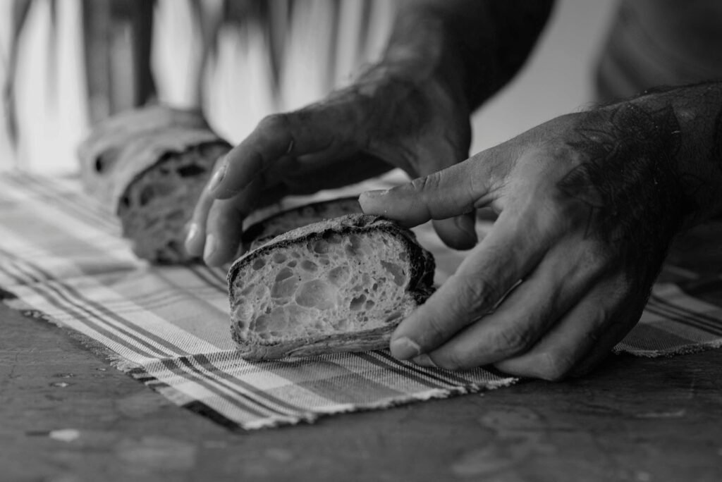 Black and white photo of hands slicing rustic artisan bread, emphasizing texture.