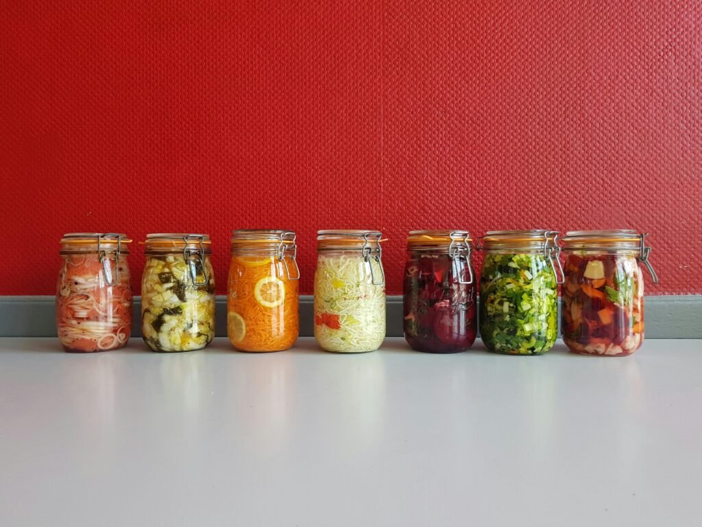 FermentarenKasa.es A row of vibrant, multicolored mason jars filled with assorted fermented vegetables against a red wall.