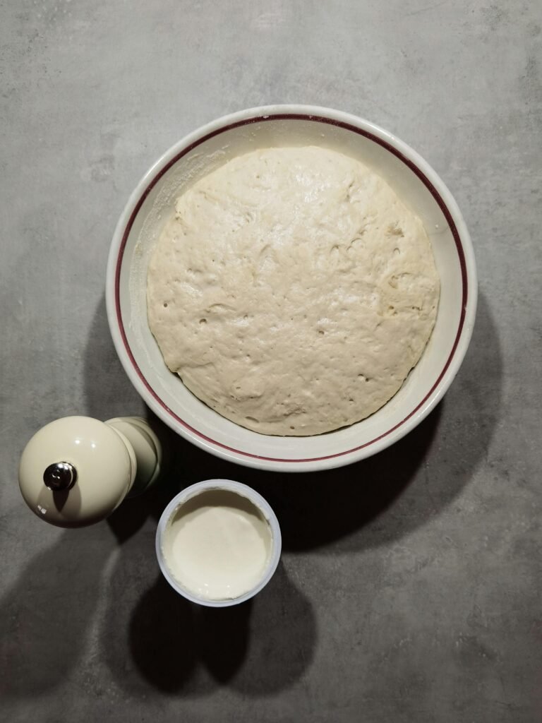 Fluffy bread dough rising in a bowl with flour and a pepper grinder nearby.
